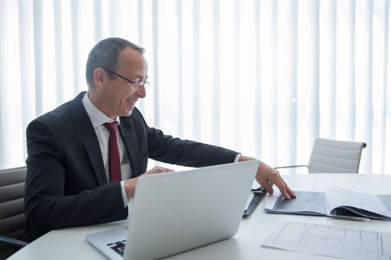 Elderly businessman in a suit working on a laptop and reviewing documents in a bright office setting.